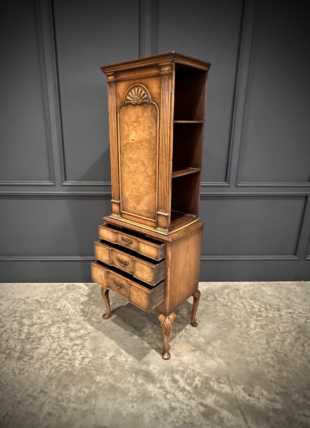 A tall, narrow Queen Anne style walnut bookcase featuring an arched burr walnut front panel, open side shelving, three drawers with brass handles, and cabriole legs with carved shell knees. View IX