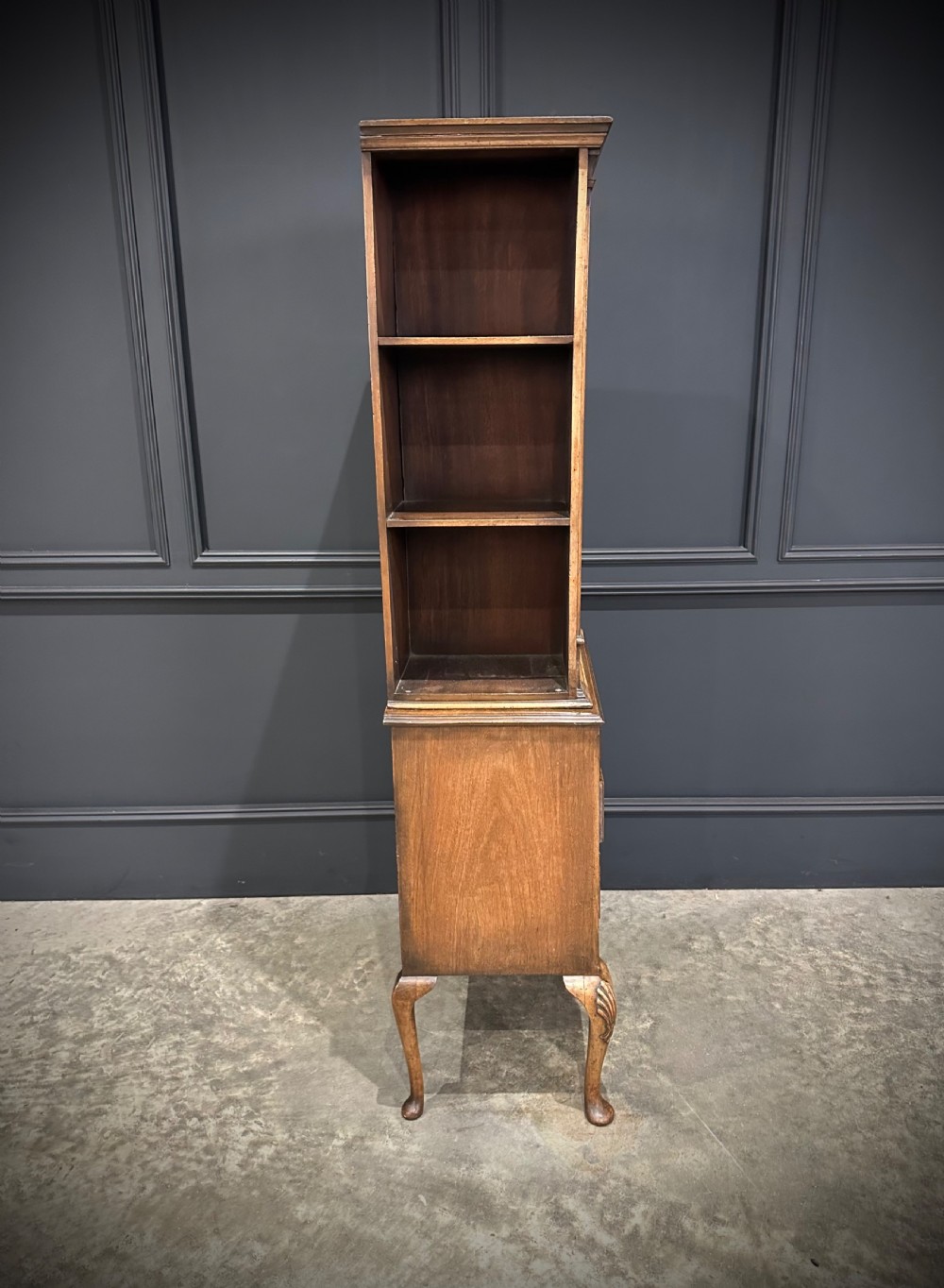 A tall, narrow Queen Anne style walnut bookcase featuring an arched burr walnut front panel, open side shelving, three drawers with brass handles, and cabriole legs with carved shell knees. View X