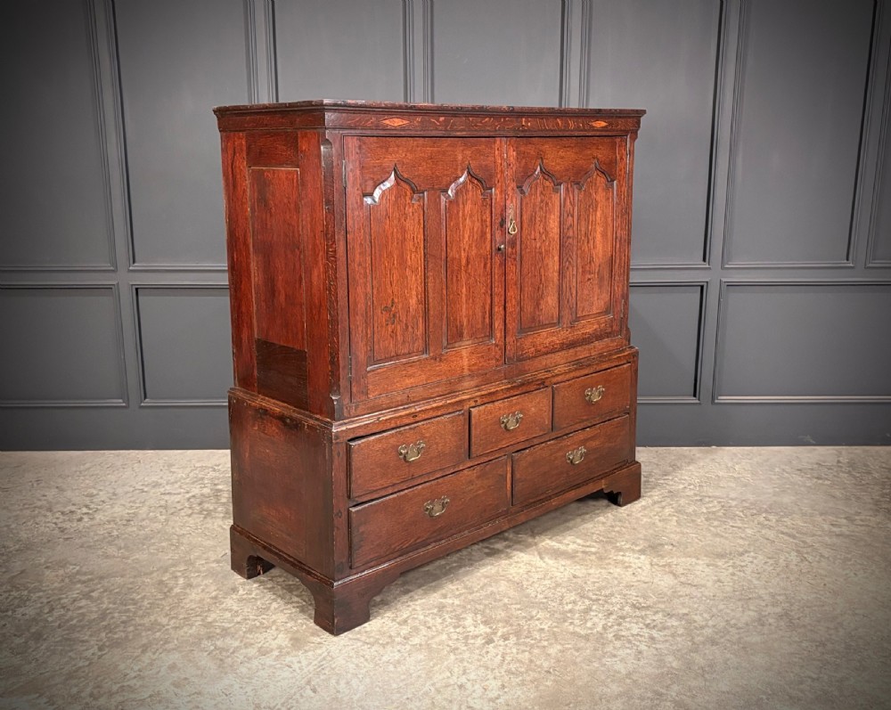 An 18th-century dwarf oak housekeepers cupboard featuring twin doors with pointed arch fielded panels over a base of five drawers.