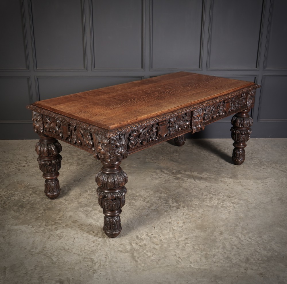 A dark carved oak writing table featuring relief frieze carvings, mask details, and turned legs. View II