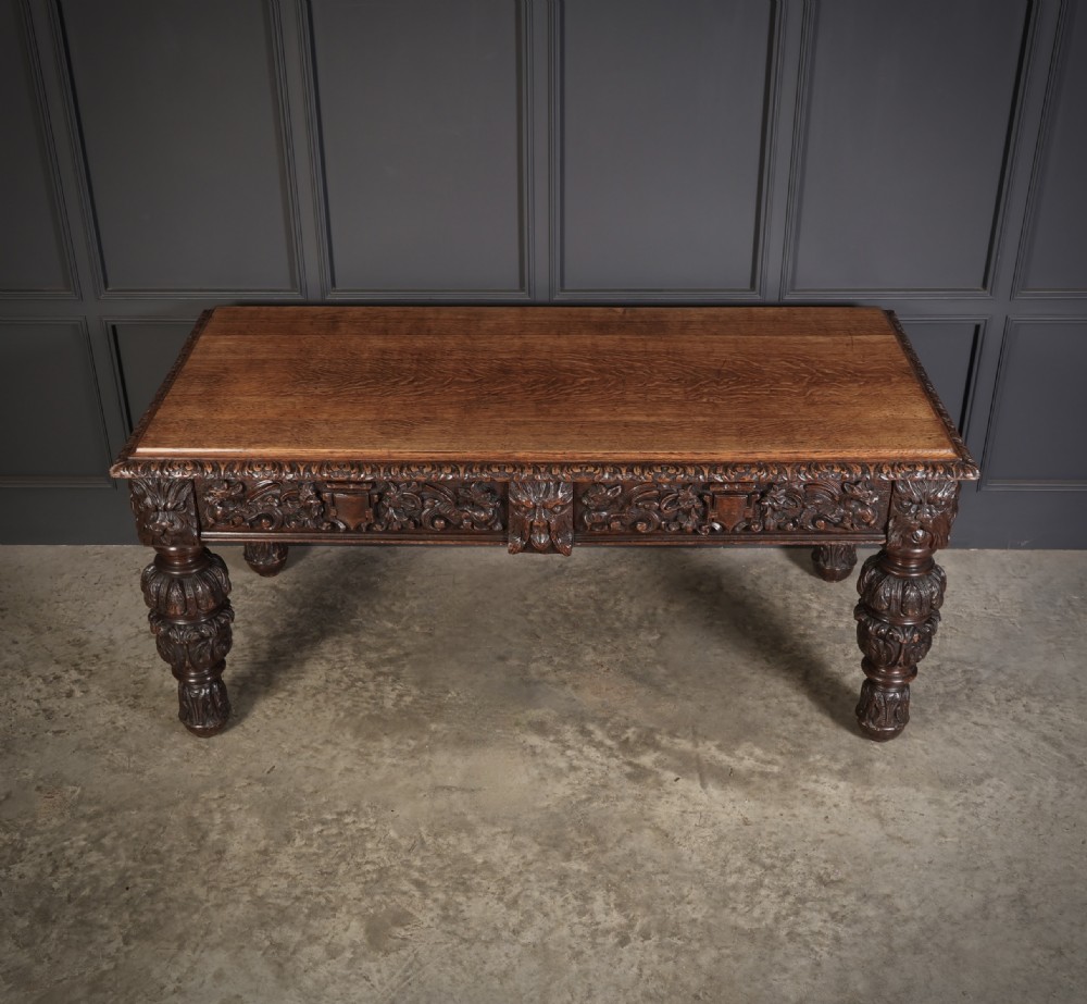 A dark carved oak writing table featuring relief frieze carvings, mask details, and turned legs. View III