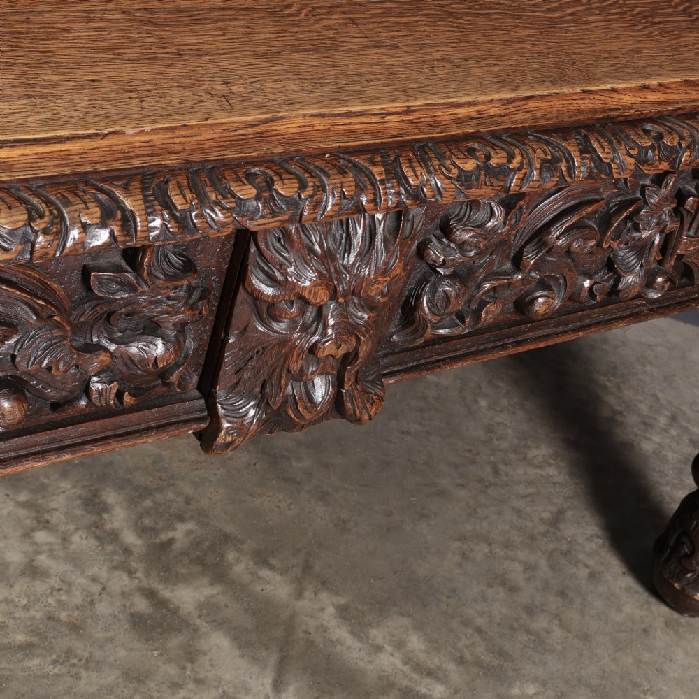 A dark carved oak writing table featuring relief frieze carvings, mask details, and turned legs. View IV