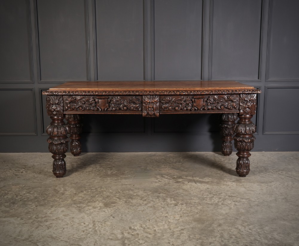 A dark carved oak writing table featuring relief frieze carvings, mask details, and turned legs. View V