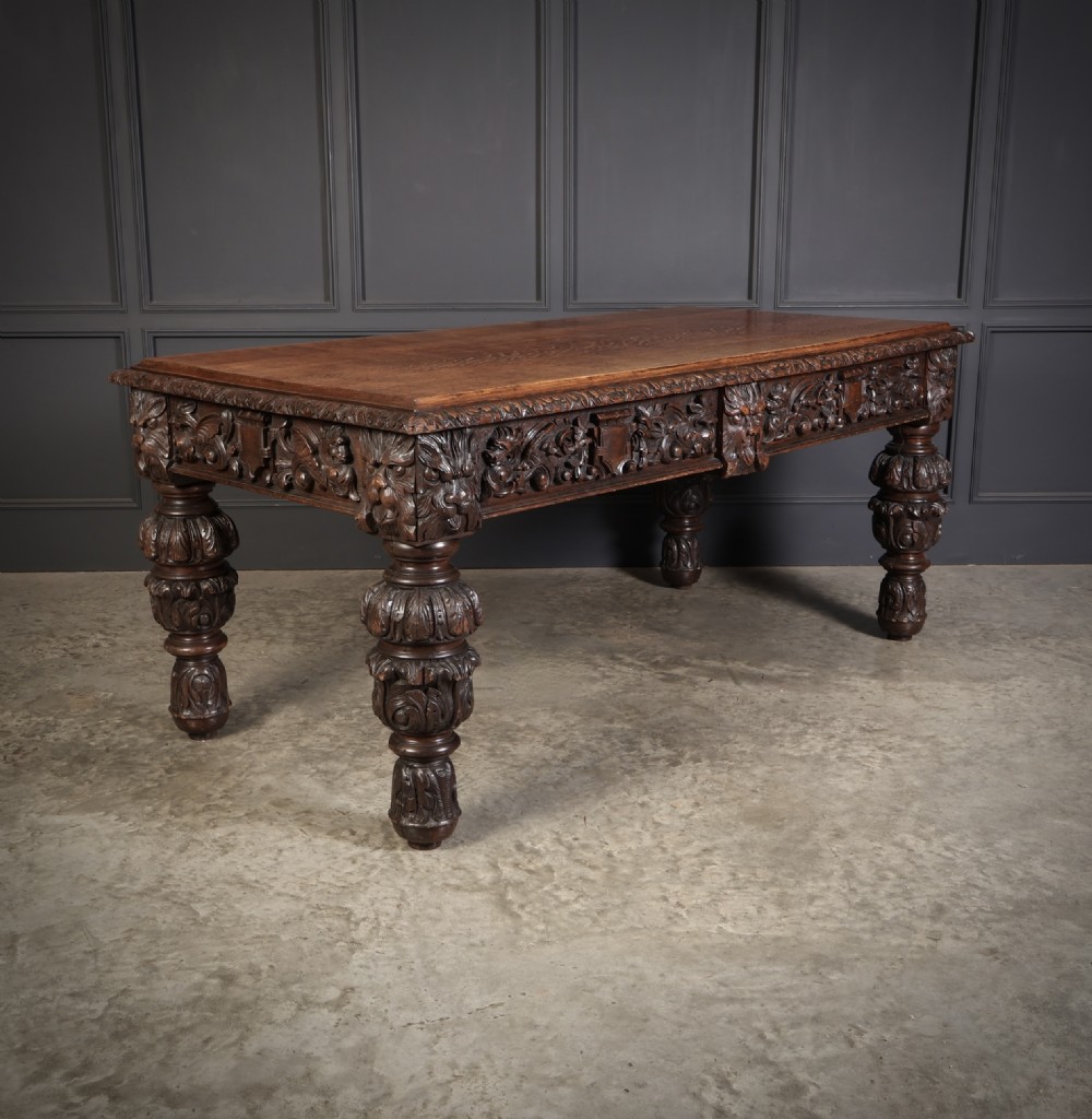 A dark carved oak writing table featuring relief frieze carvings, mask details, and turned legs.