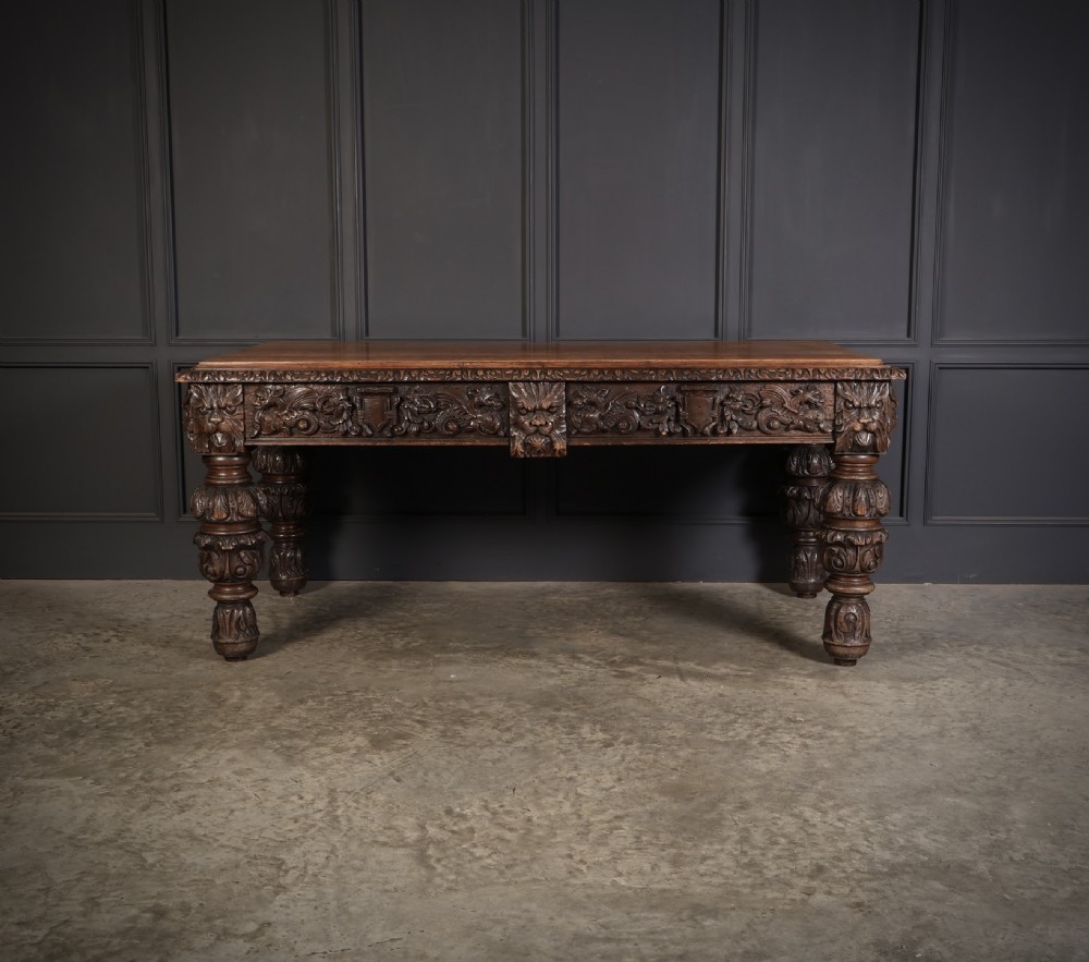 A dark carved oak writing table featuring relief frieze carvings, mask details, and turned legs. View XIV