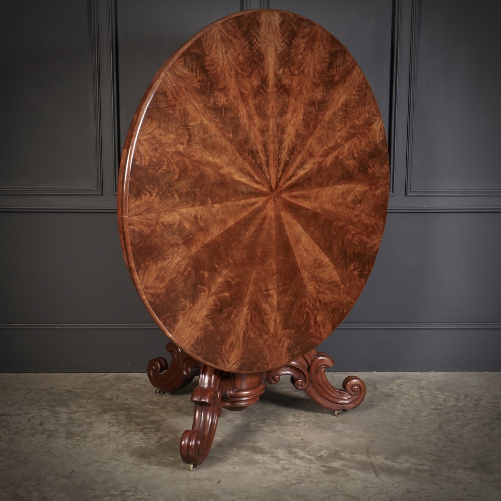A circular antique mahogany tilt-top centre table with a sunburst flame veneer top mounted on a heavy carved pedestal base with scroll feet.