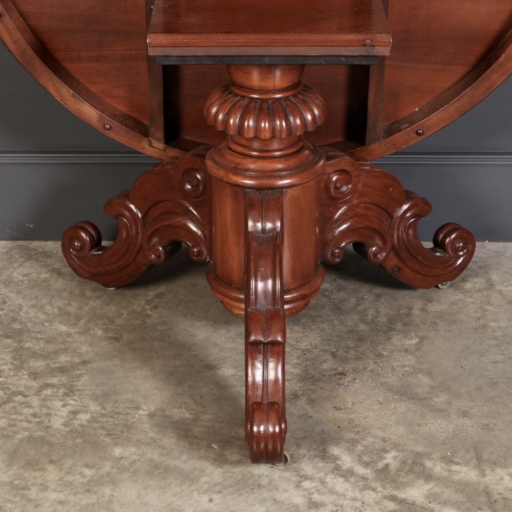A circular antique mahogany tilt-top centre table with a sunburst flame veneer top mounted on a heavy carved pedestal base with scroll feet. View II