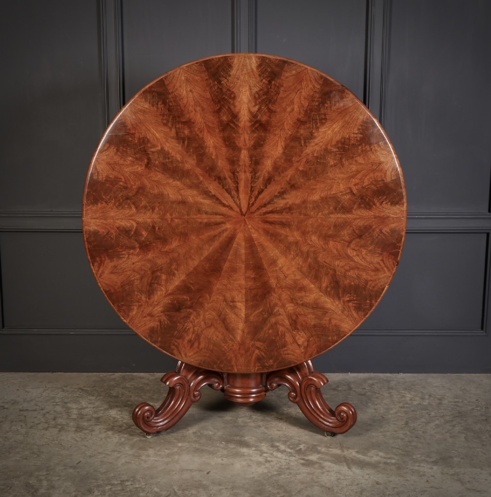 A circular antique mahogany tilt-top centre table with a sunburst flame veneer top mounted on a heavy carved pedestal base with scroll feet. View III