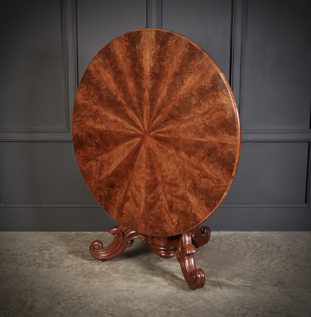 A circular antique mahogany tilt-top centre table with a sunburst flame veneer top mounted on a heavy carved pedestal base with scroll feet. View IV