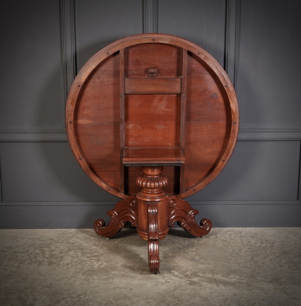 A circular antique mahogany tilt-top centre table with a sunburst flame veneer top mounted on a heavy carved pedestal base with scroll feet. View V