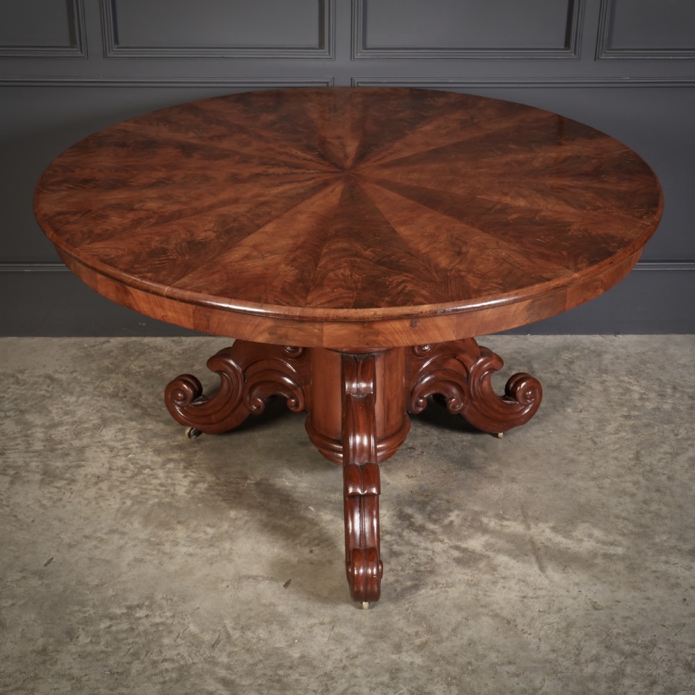 A circular antique mahogany tilt-top centre table with a sunburst flame veneer top mounted on a heavy carved pedestal base with scroll feet. View VIII