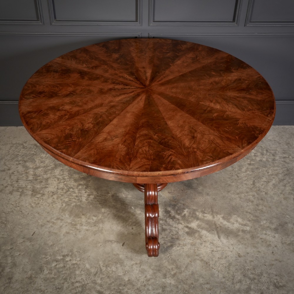 A circular antique mahogany tilt-top centre table with a sunburst flame veneer top mounted on a heavy carved pedestal base with scroll feet. View IX
