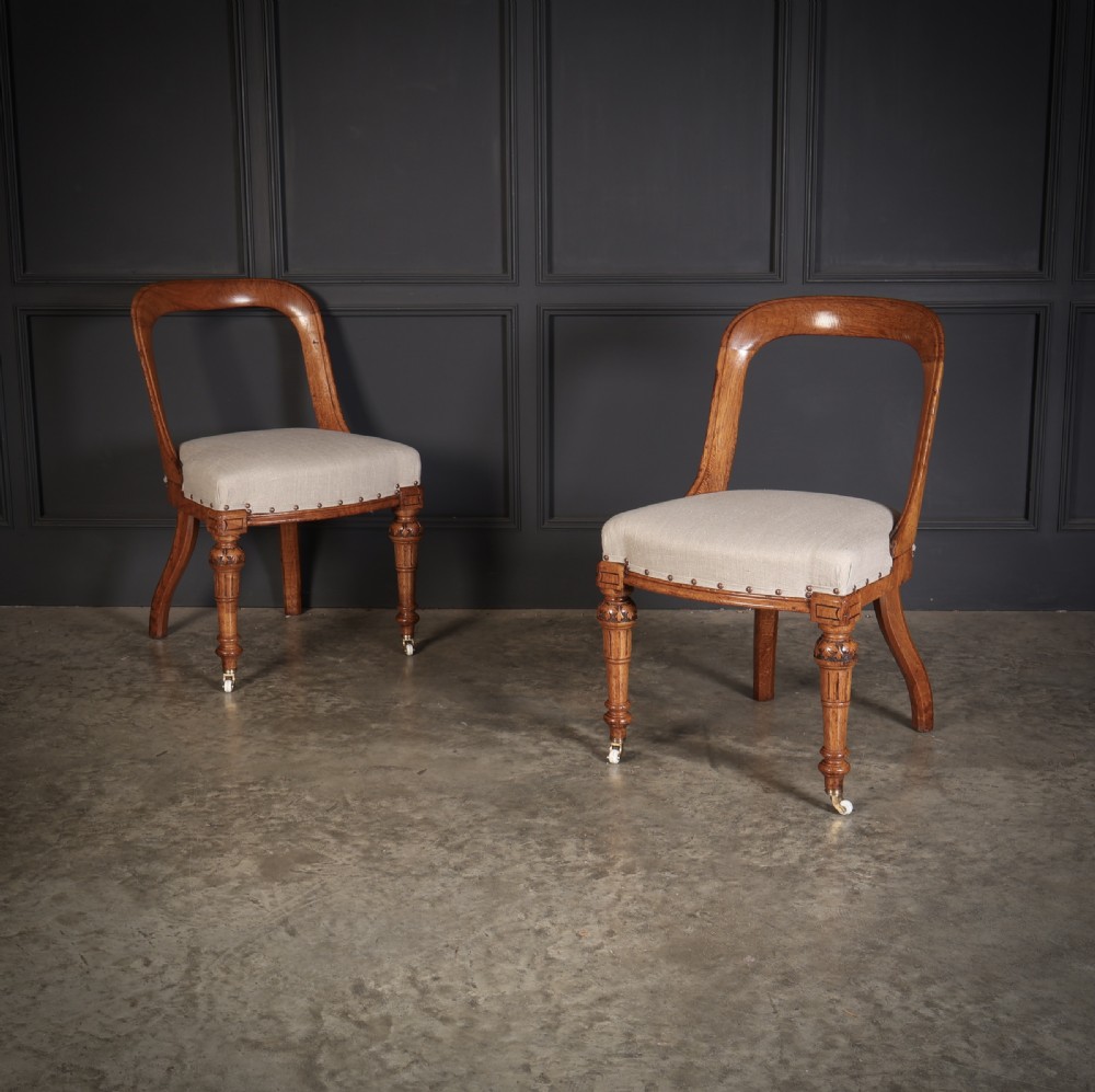 A set of ten Victorian oak dining chairs featuring curved open backs, neutral linen upholstered seats with brass studding, and turned front legs on castors. View III