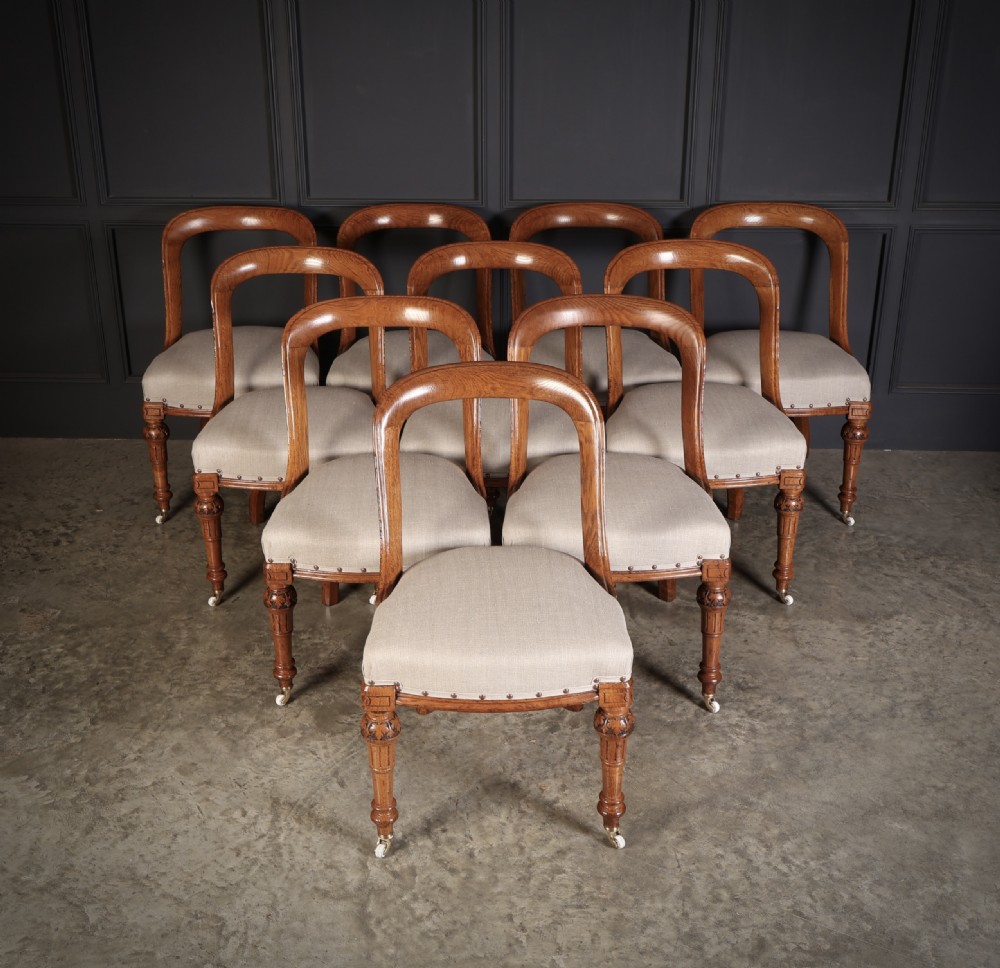 A set of ten Victorian oak dining chairs featuring curved open backs, neutral linen upholstered seats with brass studding, and turned front legs on castors. View IV