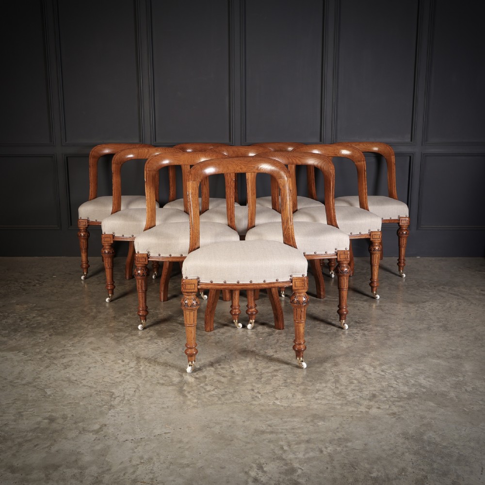 A set of ten Victorian oak dining chairs featuring curved open backs, neutral linen upholstered seats with brass studding, and turned front legs on castors.