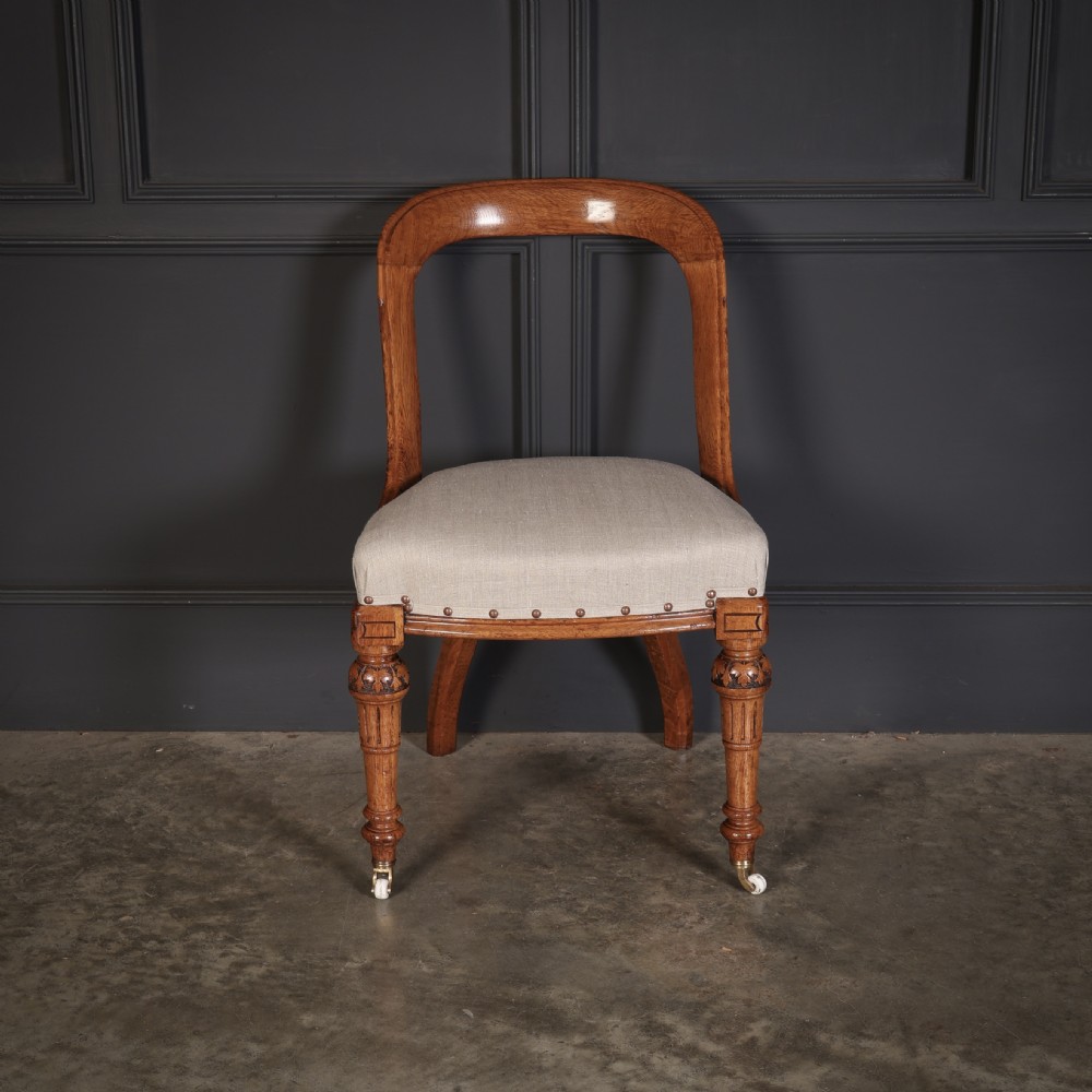 A set of ten Victorian oak dining chairs featuring curved open backs, neutral linen upholstered seats with brass studding, and turned front legs on castors. View VI