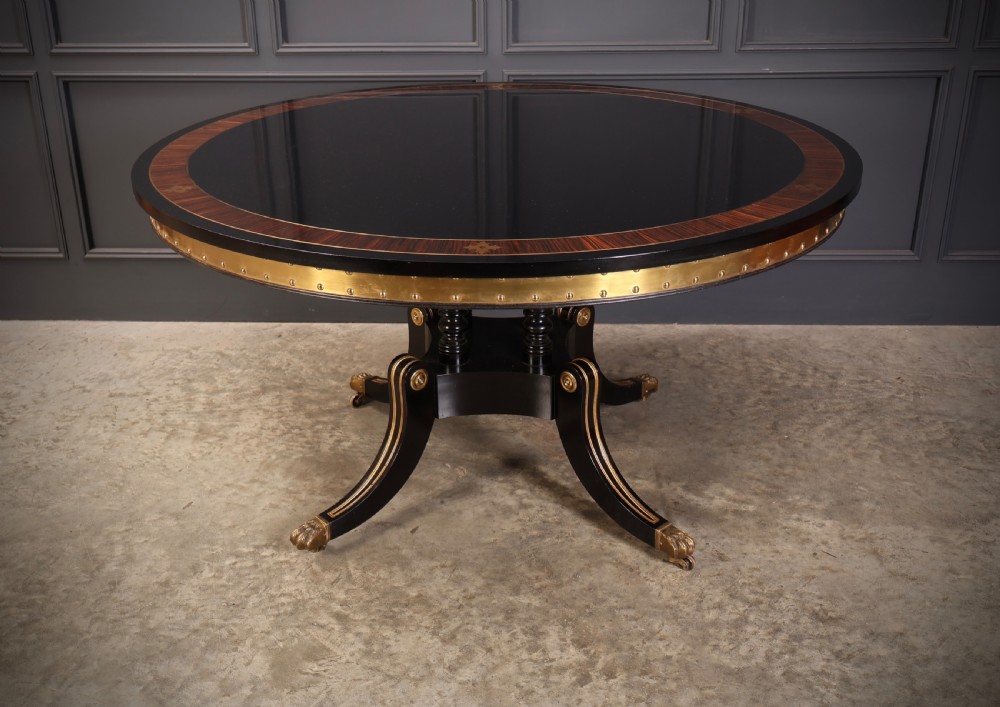 A circular ebonised centre table featuring brass line inlays, a crossbanded top border, a brass apron rim, and a quadruped pedestal base with brass lion paw feet.