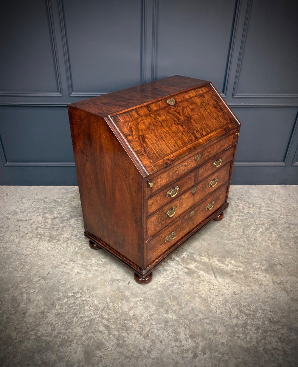 An antique Queen Anne walnut bureau featuring a sloping write-down front above drawers with brass handles, raised on compressed bun feet.
