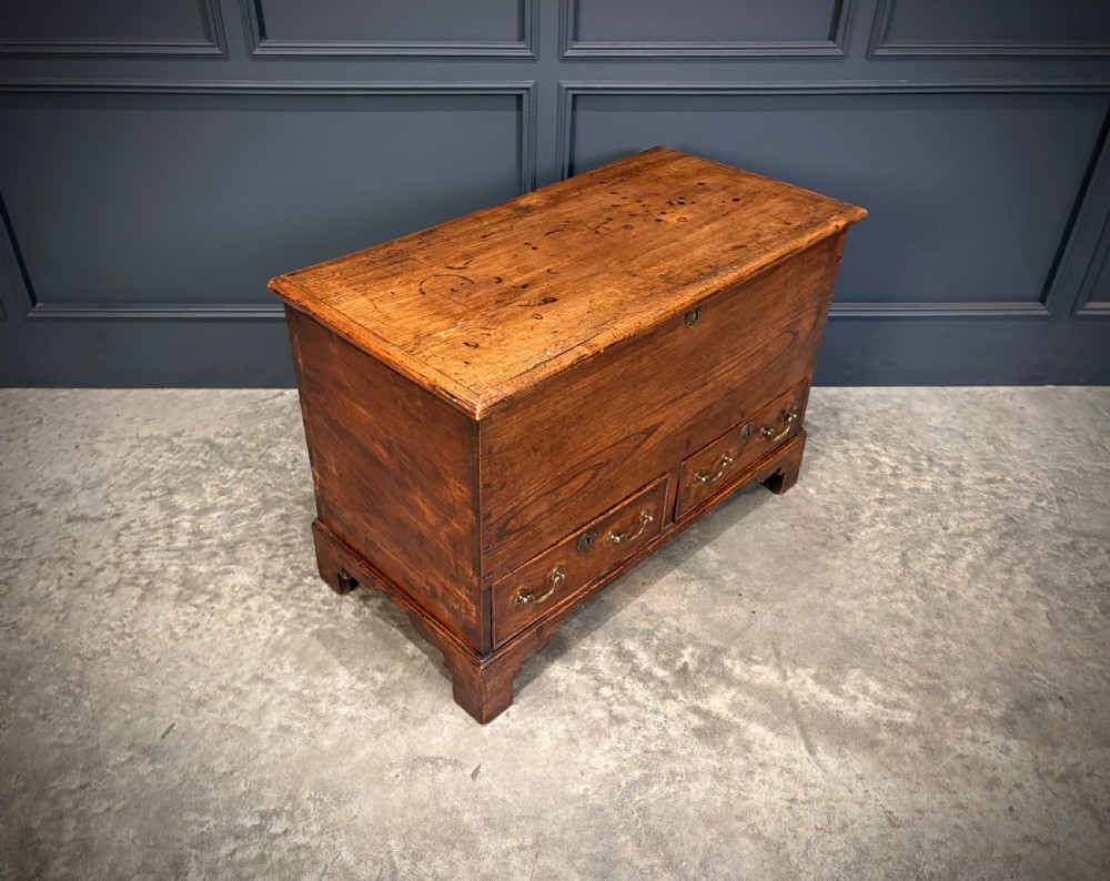 A Georgian elm blanket box featuring a hinged top lid, two lower drawers with brass swan-neck handles, and shaped bracket feet. View II