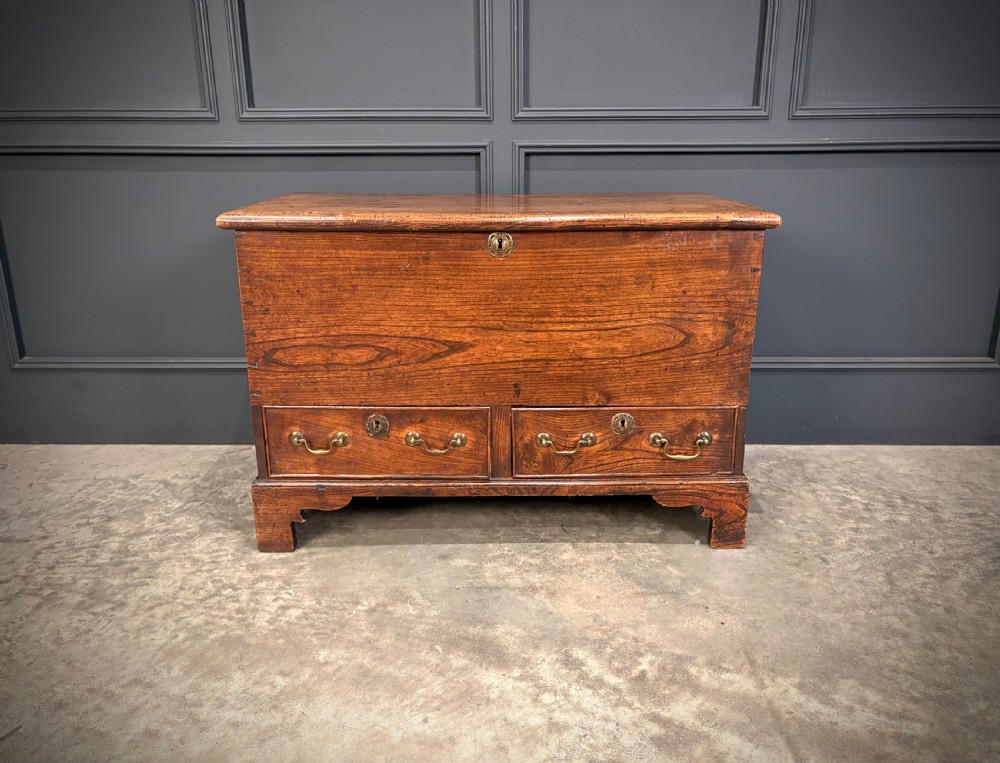 A Georgian elm blanket box featuring a hinged top lid, two lower drawers with brass swan-neck handles, and shaped bracket feet. View V