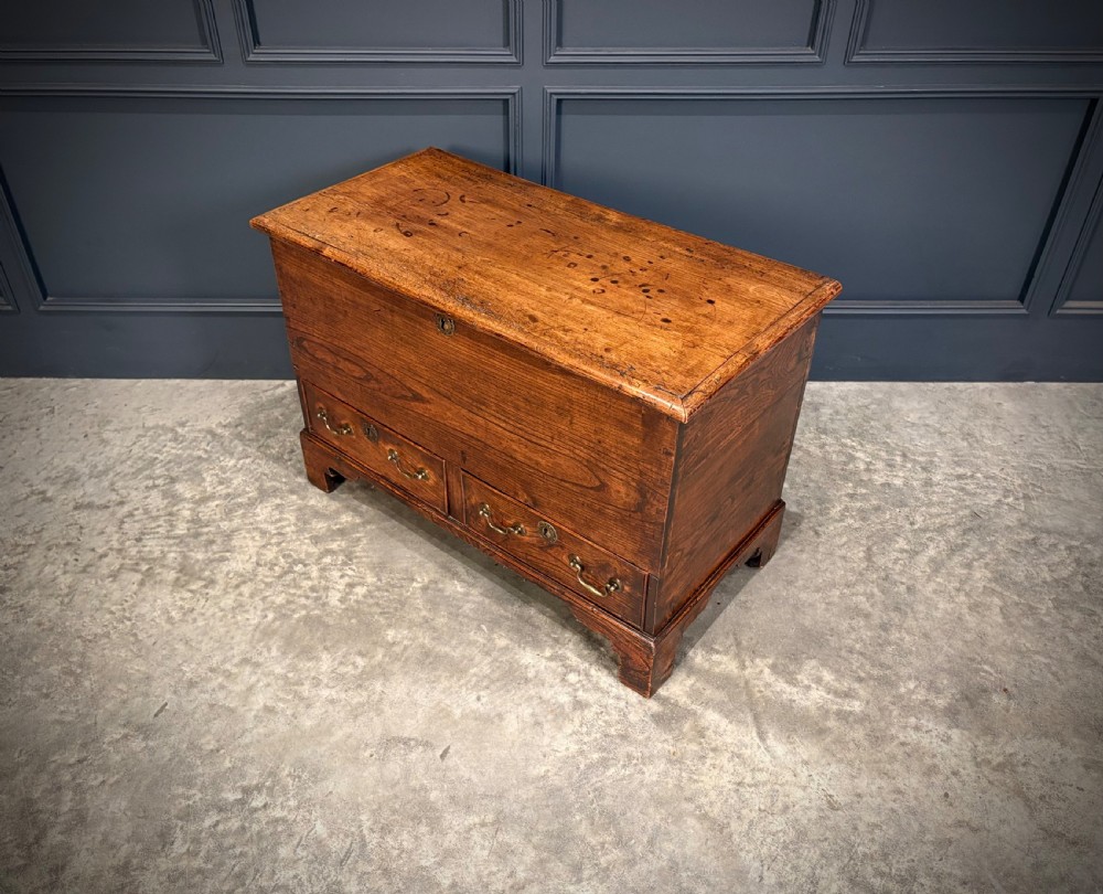 A Georgian elm blanket box featuring a hinged top lid, two lower drawers with brass swan-neck handles, and shaped bracket feet. View VI