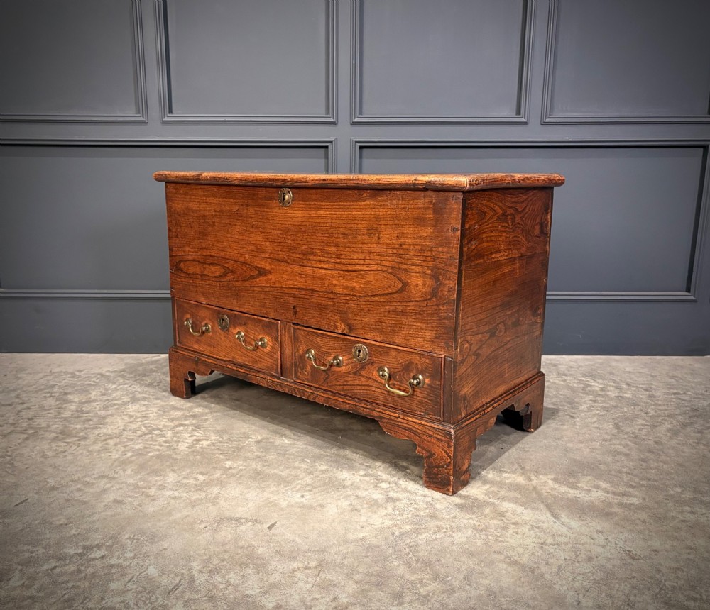 A Georgian elm blanket box featuring a hinged top lid, two lower drawers with brass swan-neck handles, and shaped bracket feet.