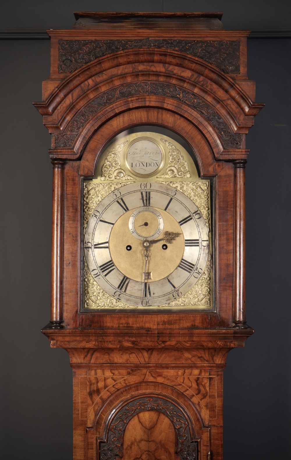 An 18th-century carved walnut longcase clock featuring an arched hood, brass dial with silvered chapter ring, and decorative carved central door panel. View II
