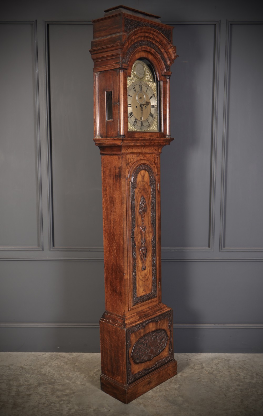 An 18th-century carved walnut longcase clock featuring an arched hood, brass dial with silvered chapter ring, and decorative carved central door panel. View V