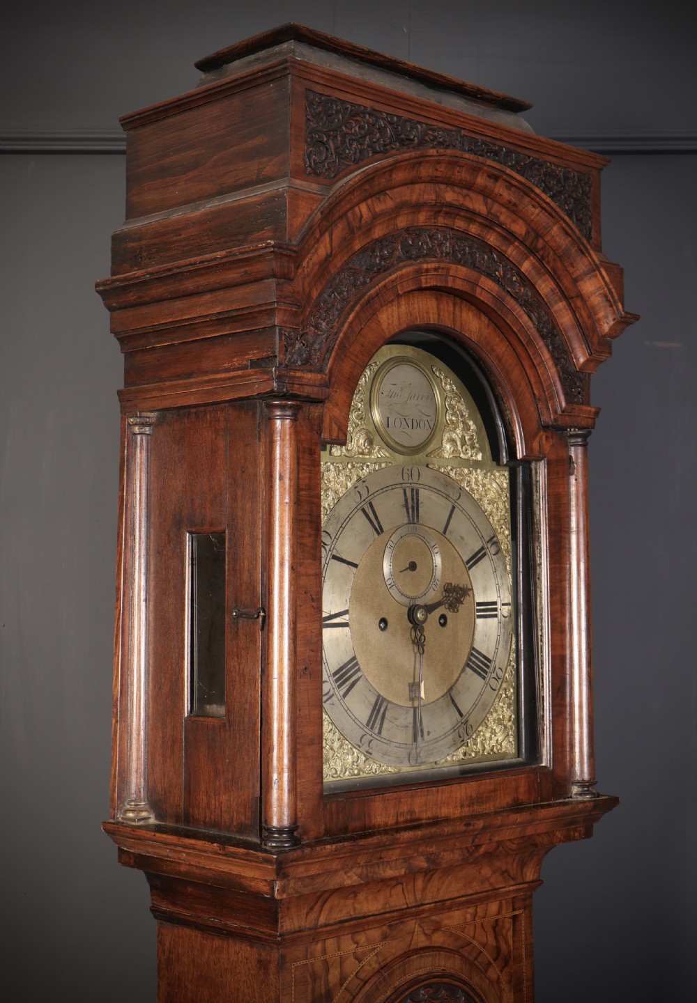 An 18th-century carved walnut longcase clock featuring an arched hood, brass dial with silvered chapter ring, and decorative carved central door panel. View VI
