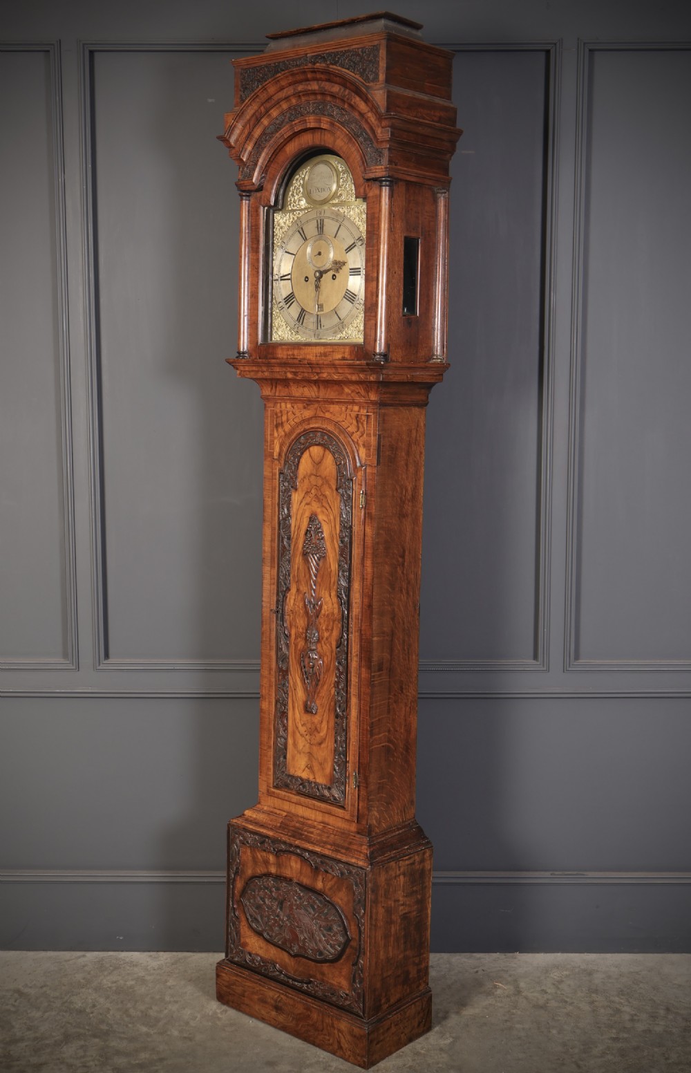 An 18th-century carved walnut longcase clock featuring an arched hood, brass dial with silvered chapter ring, and decorative carved central door panel. View VII