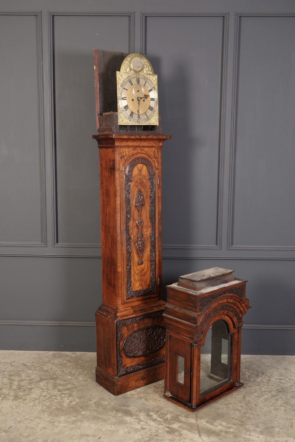 An 18th-century carved walnut longcase clock featuring an arched hood, brass dial with silvered chapter ring, and decorative carved central door panel. View XII