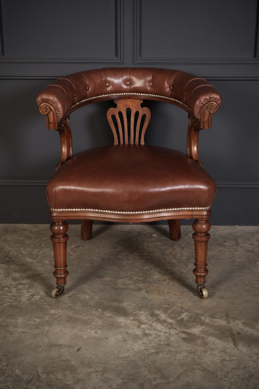 A pair of Victorian mahogany captains desk chairs featuring button-tufted brown leather horseshoe armrests, padded leather seats with studding, turned front legs, and brass castors. View V