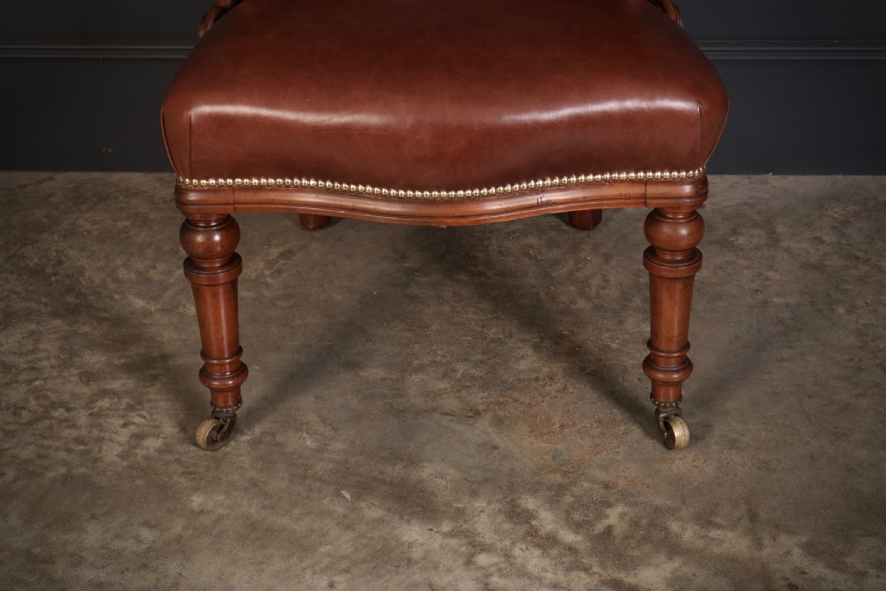 A pair of Victorian mahogany captains desk chairs featuring button-tufted brown leather horseshoe armrests, padded leather seats with studding, turned front legs, and brass castors. View VI