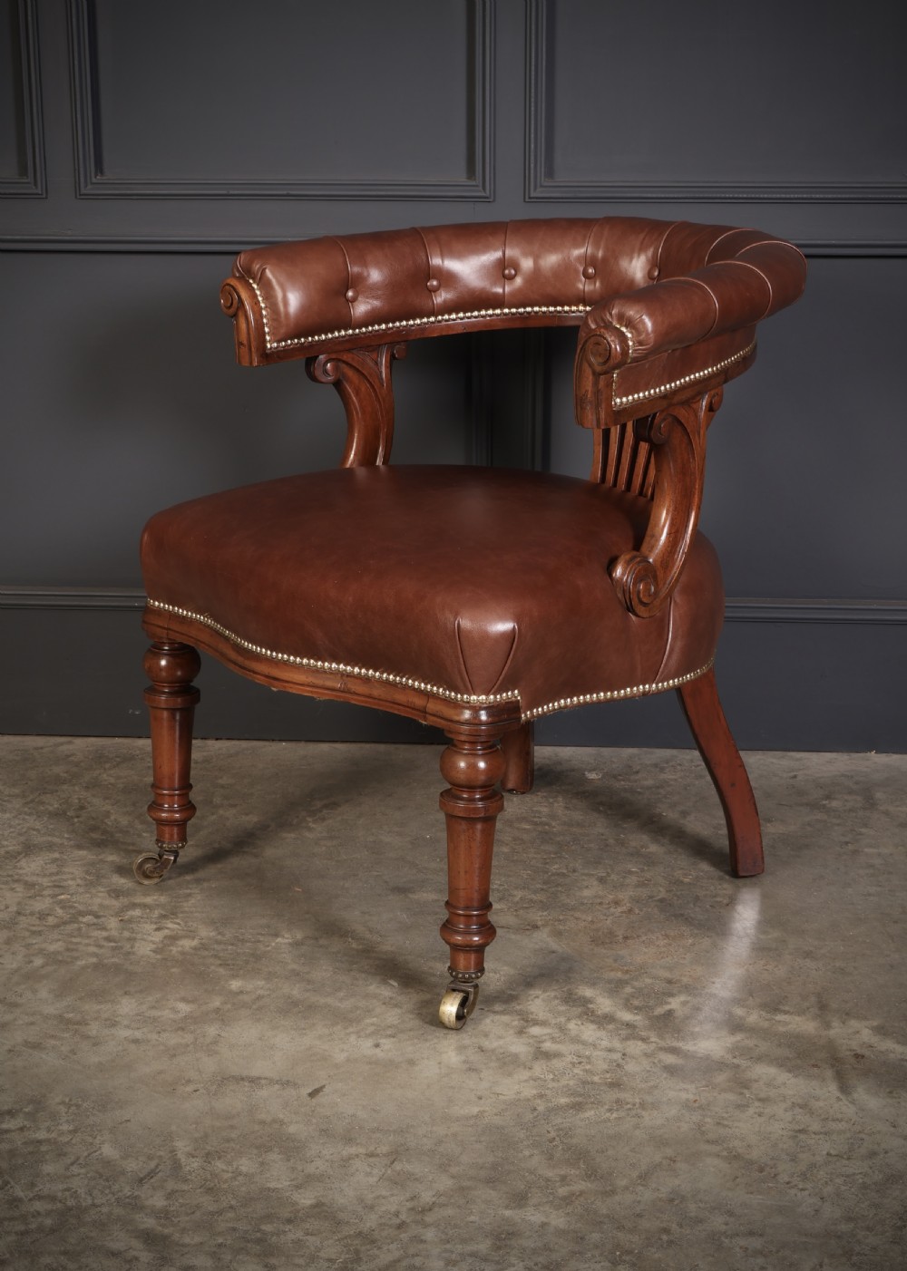 A pair of Victorian mahogany captains desk chairs featuring button-tufted brown leather horseshoe armrests, padded leather seats with studding, turned front legs, and brass castors. View VIII
