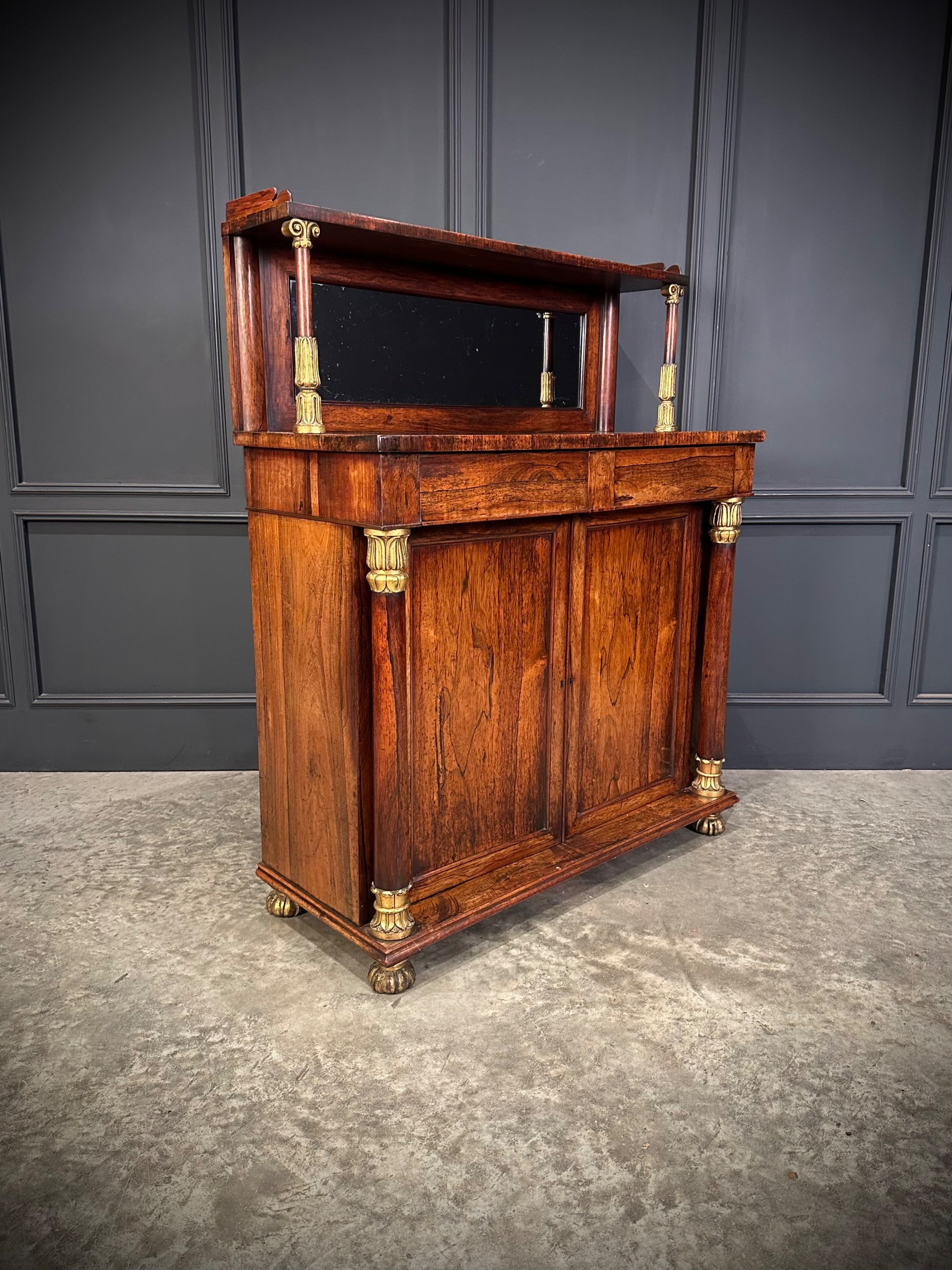 A William IV dark rosewood chiffonier featuring a mirrored top shelf supported by gilded columns over a two-door cabinet with paw feet.