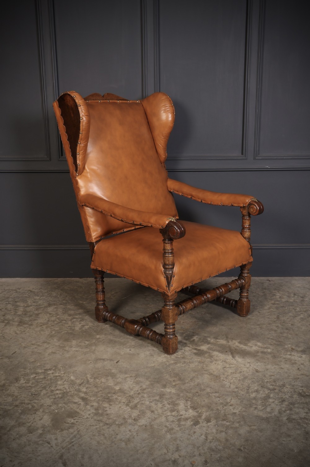 A nineteenth-century wingback armchair featuring tan leather upholstery with brass stud detailing, carved oak scroll armrests, and turned legs with connecting stretchers.
