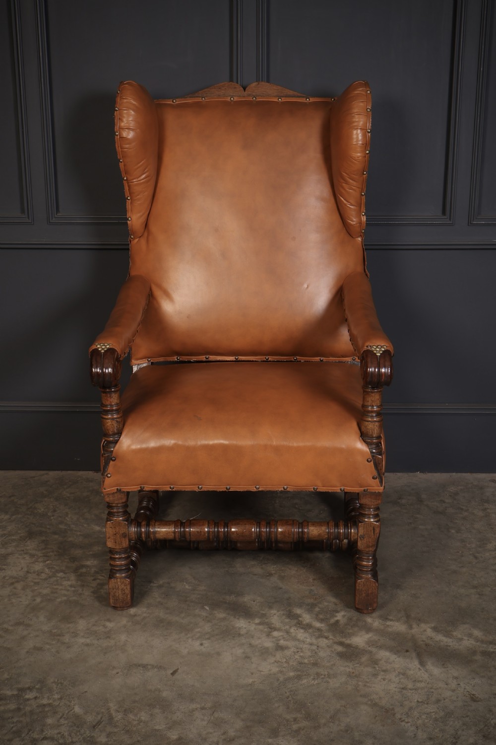 A nineteenth-century wingback armchair featuring tan leather upholstery with brass stud detailing, carved oak scroll armrests, and turned legs with connecting stretchers. View II