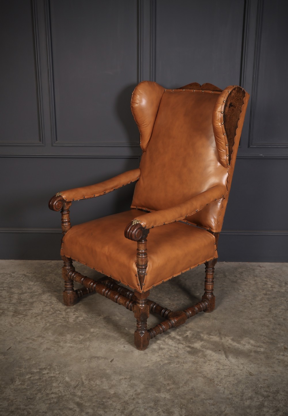 A nineteenth-century wingback armchair featuring tan leather upholstery with brass stud detailing, carved oak scroll armrests, and turned legs with connecting stretchers. View III