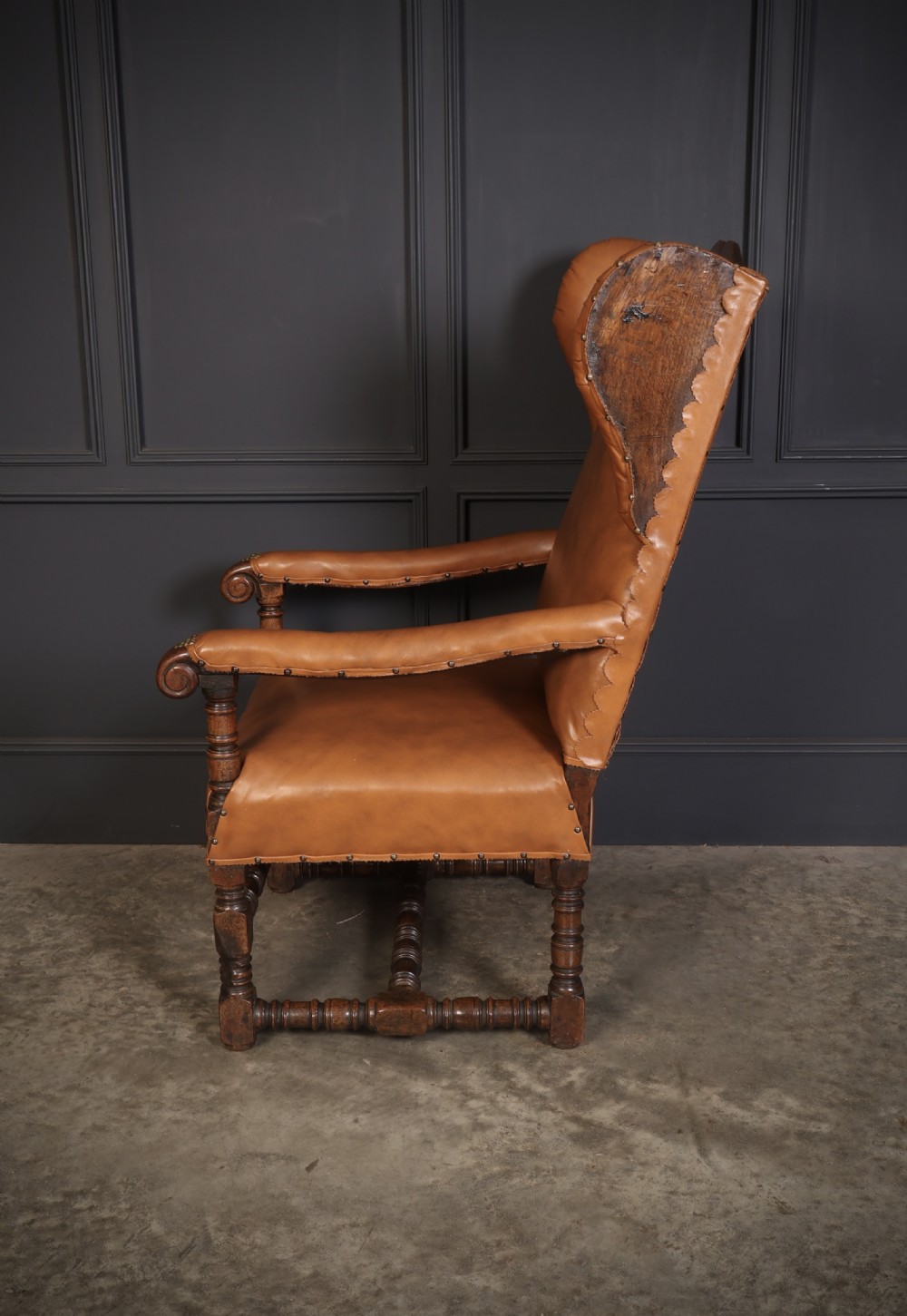 A nineteenth-century wingback armchair featuring tan leather upholstery with brass stud detailing, carved oak scroll armrests, and turned legs with connecting stretchers. View V