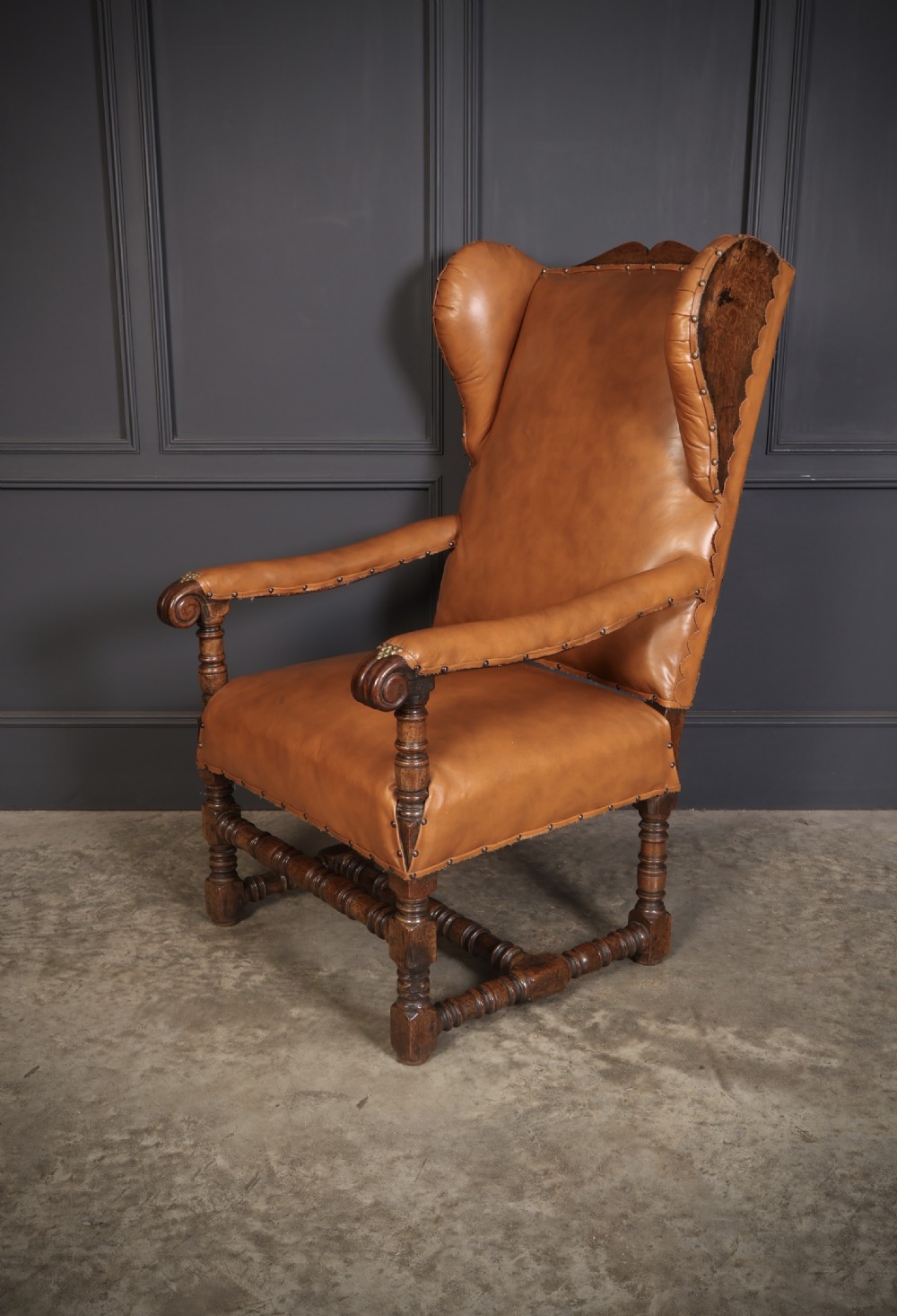 A nineteenth-century wingback armchair featuring tan leather upholstery with brass stud detailing, carved oak scroll armrests, and turned legs with connecting stretchers. View VI