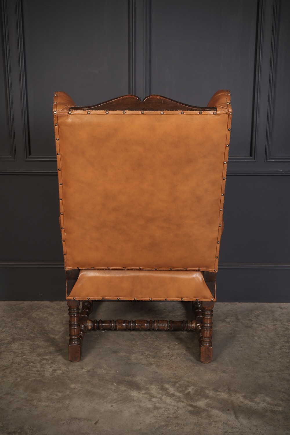 A nineteenth-century wingback armchair featuring tan leather upholstery with brass stud detailing, carved oak scroll armrests, and turned legs with connecting stretchers. View VII