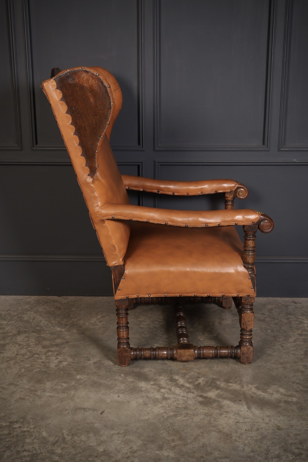 A nineteenth-century wingback armchair featuring tan leather upholstery with brass stud detailing, carved oak scroll armrests, and turned legs with connecting stretchers. View VIII