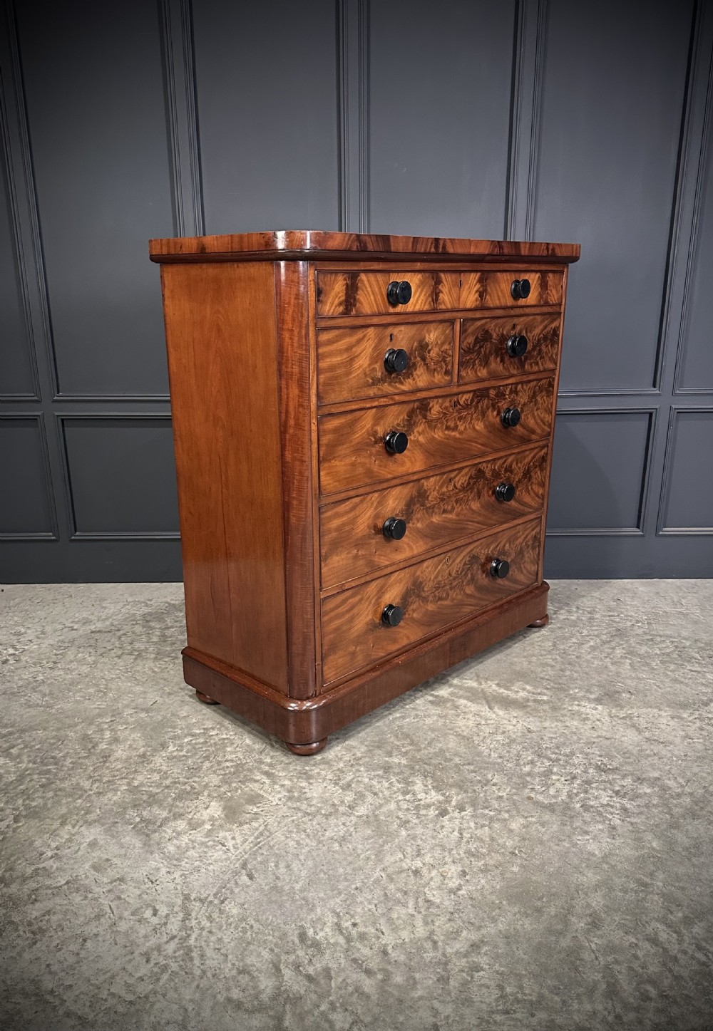 A tall Victorian flame mahogany chest of drawers featuring a decorative top drawer, five lower drawers with dark turned handles, rounded pilasters, and a solid plinth base.