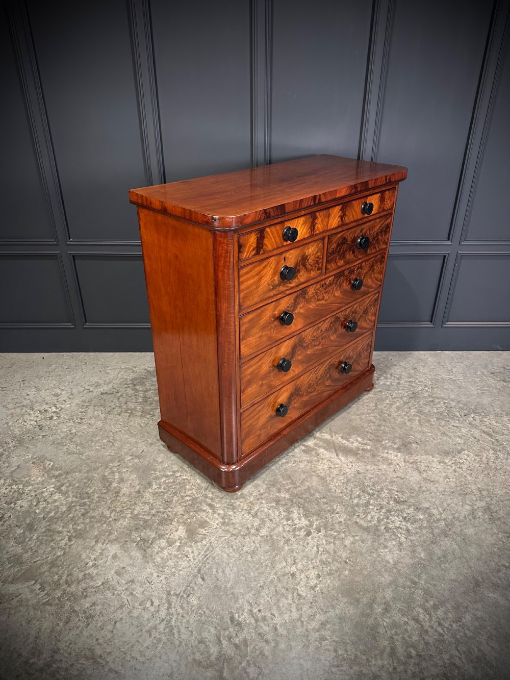 A tall Victorian flame mahogany chest of drawers featuring a decorative top drawer, five lower drawers with dark turned handles, rounded pilasters, and a solid plinth base. View II