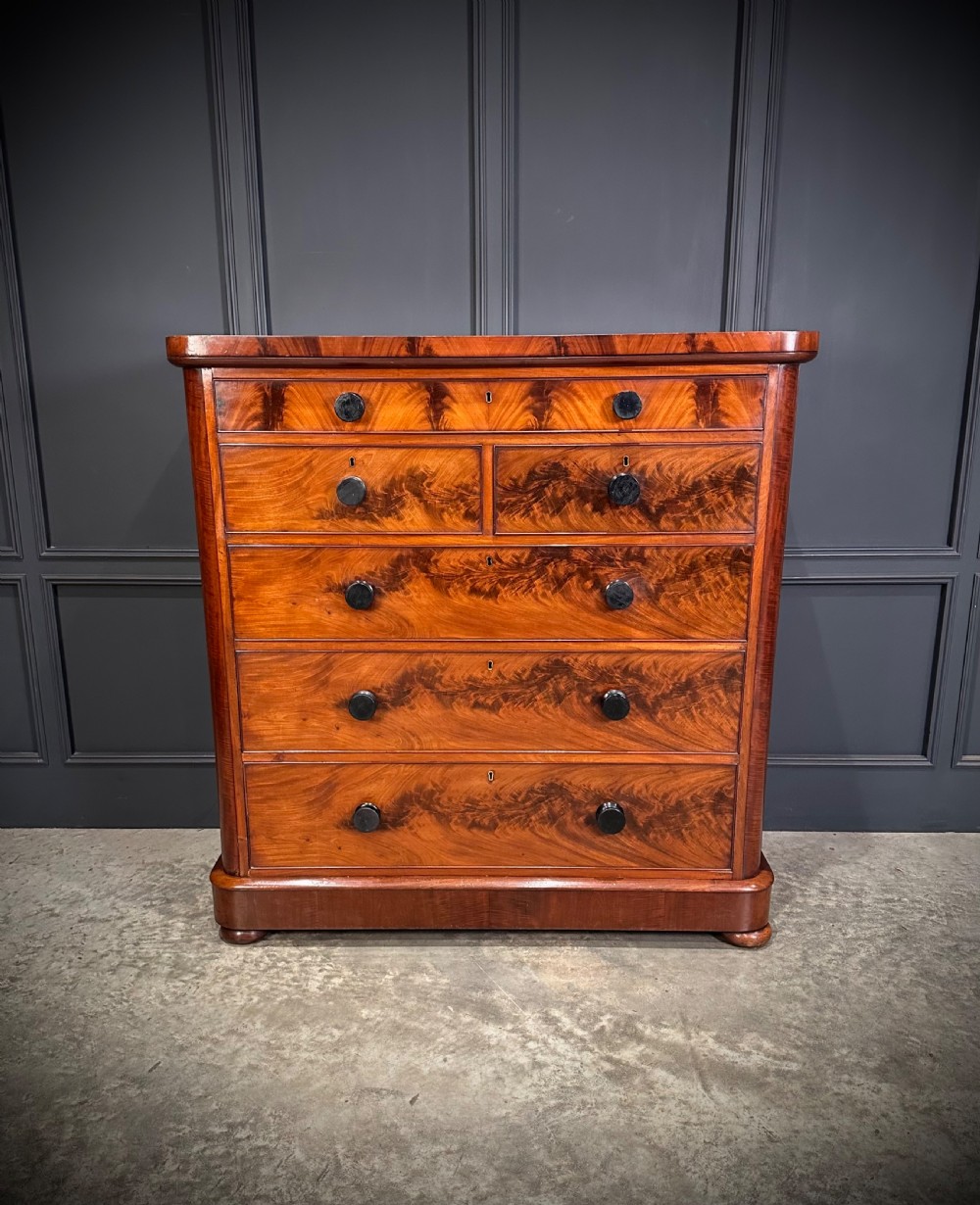 A tall Victorian flame mahogany chest of drawers featuring a decorative top drawer, five lower drawers with dark turned handles, rounded pilasters, and a solid plinth base. View III