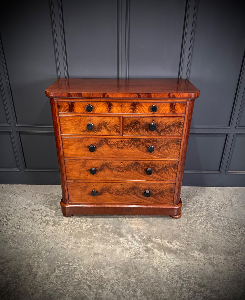 A tall Victorian flame mahogany chest of drawers featuring a decorative top drawer, five lower drawers with dark turned handles, rounded pilasters, and a solid plinth base. View IV