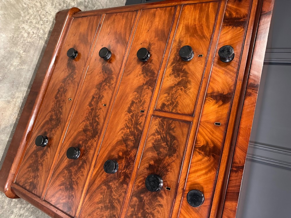 A tall Victorian flame mahogany chest of drawers featuring a decorative top drawer, five lower drawers with dark turned handles, rounded pilasters, and a solid plinth base. View V