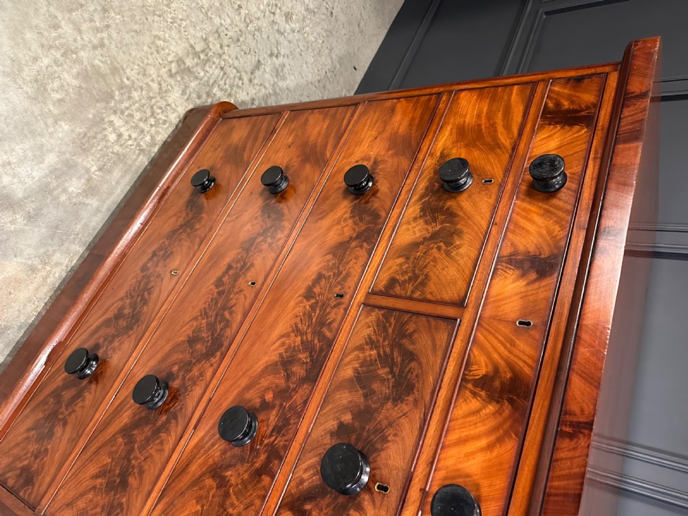 A tall Victorian flame mahogany chest of drawers featuring a decorative top drawer, five lower drawers with dark turned handles, rounded pilasters, and a solid plinth base. View VI
