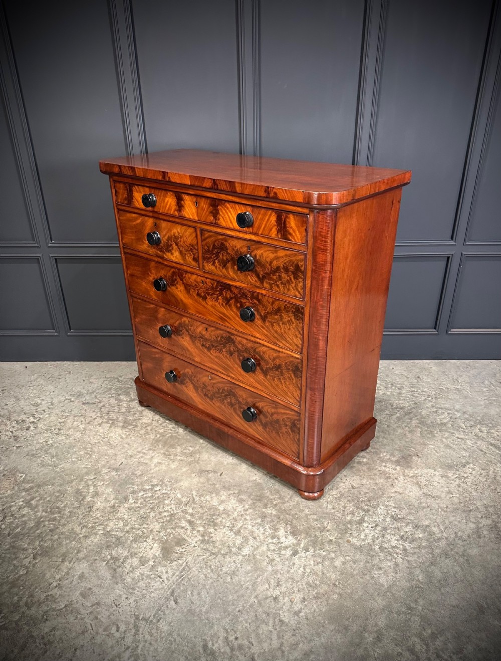 A tall Victorian flame mahogany chest of drawers featuring a decorative top drawer, five lower drawers with dark turned handles, rounded pilasters, and a solid plinth base. View VII