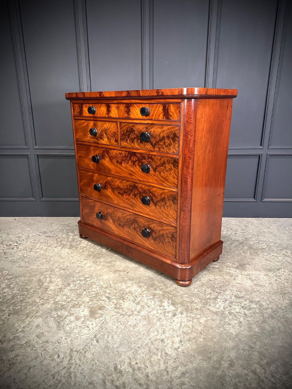 A tall Victorian flame mahogany chest of drawers featuring a decorative top drawer, five lower drawers with dark turned handles, rounded pilasters, and a solid plinth base. View VIII